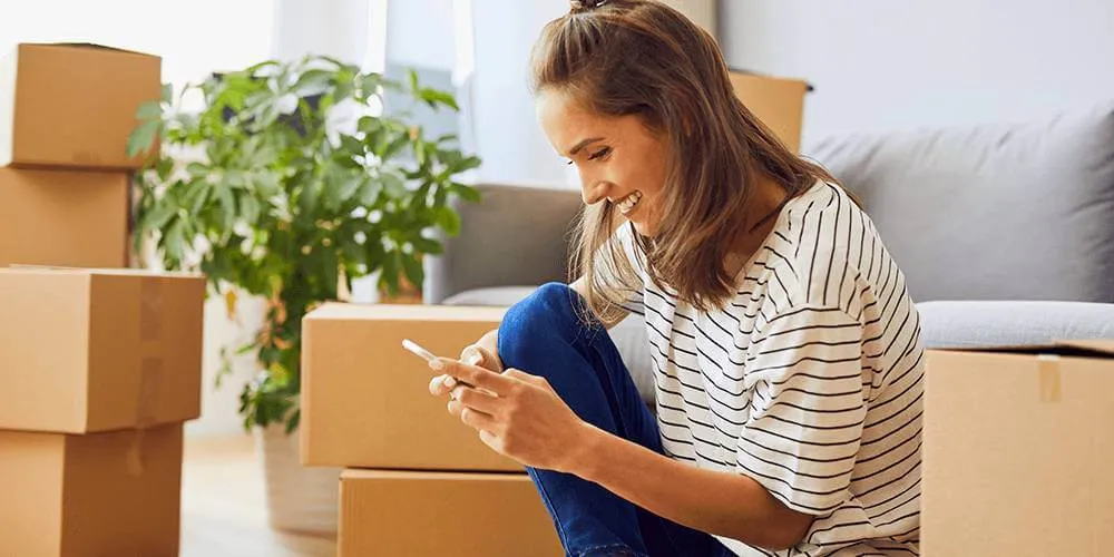 Woman sat smiling at her phone in her living room with moving boxes surrounding her