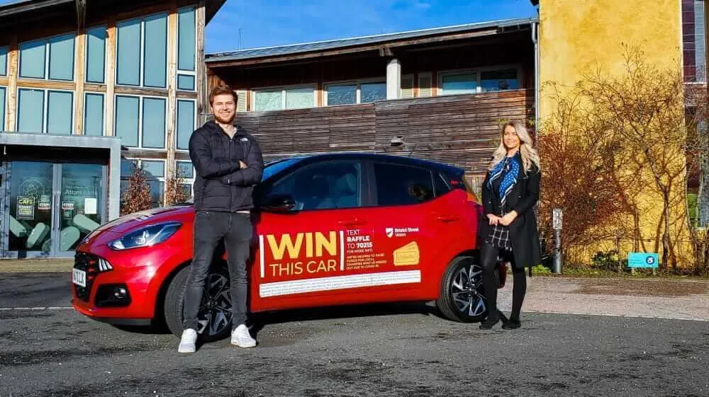 Two people standing in front of a car that is available to win in the raffle