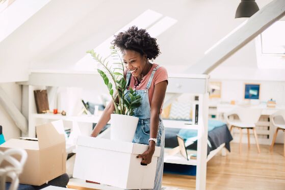 Very excited woman wearing dungarees, holding a white moving box with a potted plant on top of it