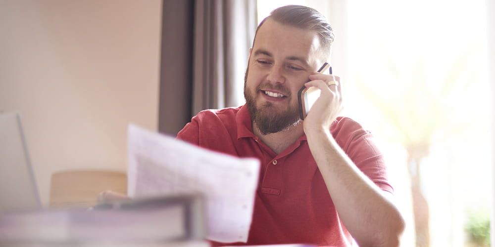Bearded man with red polo shirt is sat down on the phone whilst smiling and looking at a piece of paper