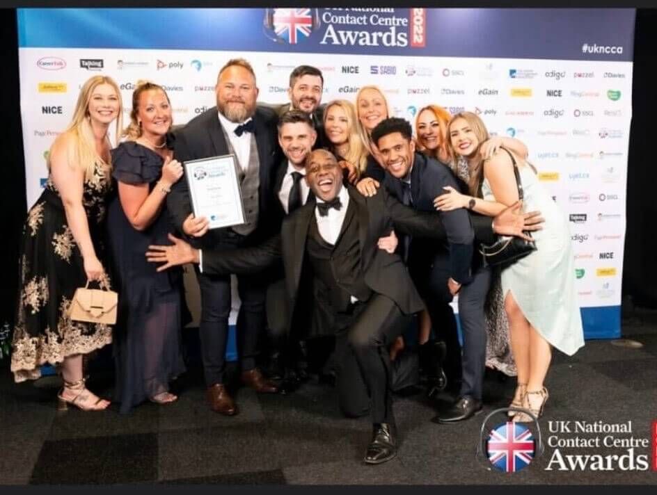 The Atom bank Contact centre team holding an award they won at the UK National Contact Centre awards with Ainsley Harriott crouching and smiling in front of them