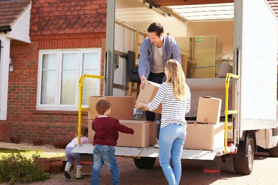 Family of four unpacking a moving truck. A man is stood in truck passing boxes down to the woman whilst children walk around the garden
