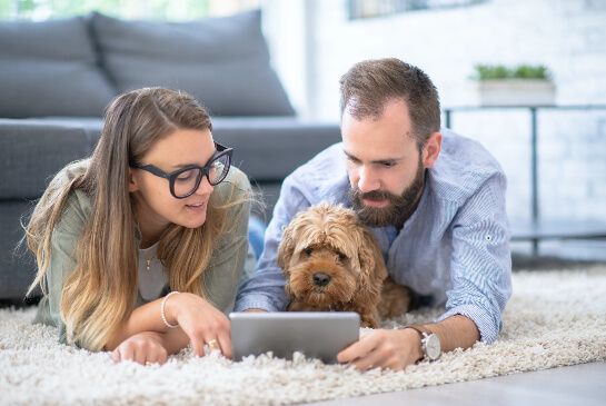 Man, woman and dog lying together on the floor of a living room looking at an ipad togther