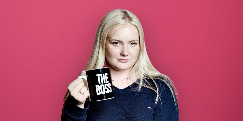 A picture of a woman infront of a pinkish background holding a mug which has the phrase 'The Boss' printed on it