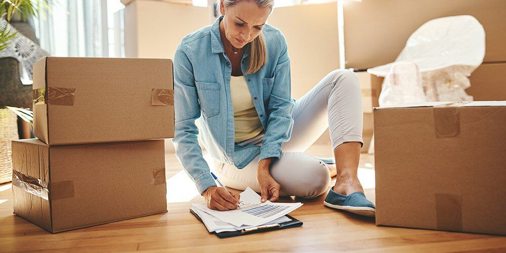 Woman sat on the floor among cardboard boxes, looking down at a moving home checklist and ticking off a completed task