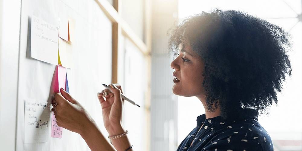 Woman looking at post it notes on a white board. She is focusing on a pink one she is holding and is about to write on it