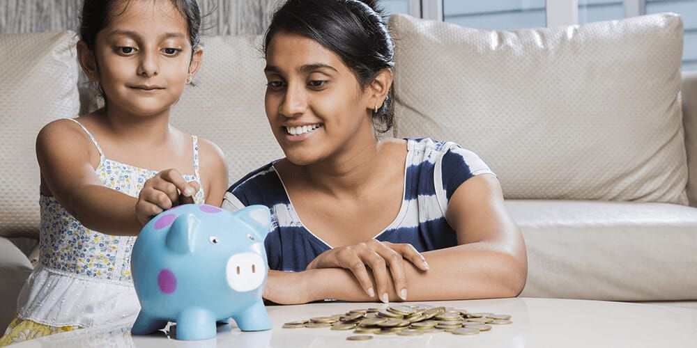 Mother and daughter sat on the floor next to a coffee table with a small pile of coins and a blue and purple piggy bank on top. The girl is dropping a coin into the piggy bank whilst her mother smiles