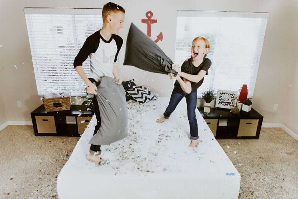 A brother and his younger sister are pillow fighting on a bed. There are feathers covering the room as the sister goes to hit her brother