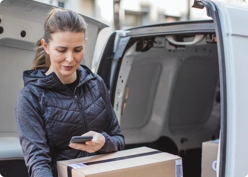 Business owner checking her phone whilst unpacking boxes from the back of a van