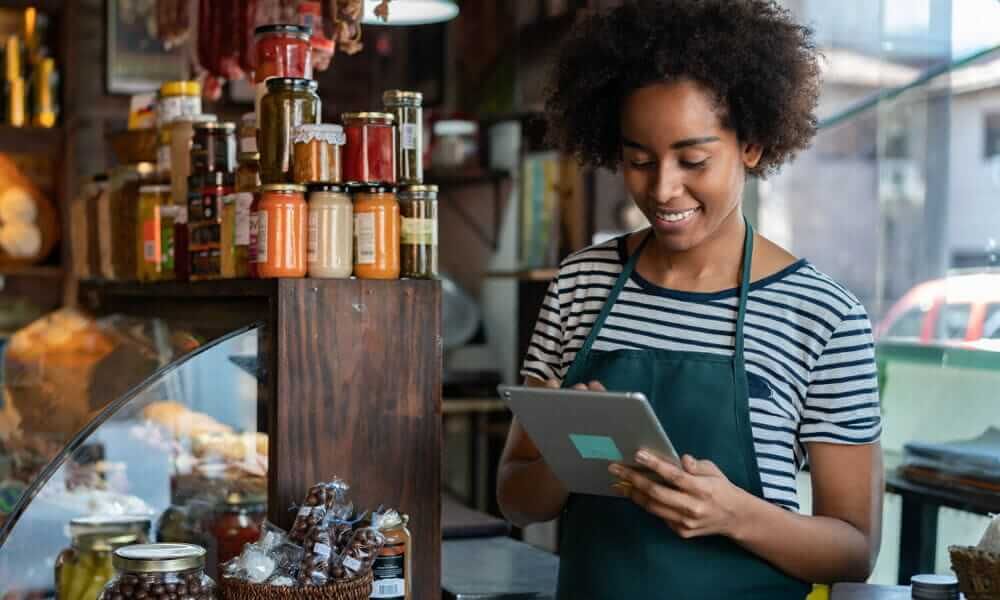 Woman wearing a green apron smiling at her ipad whilst stood in a deli next to some jams