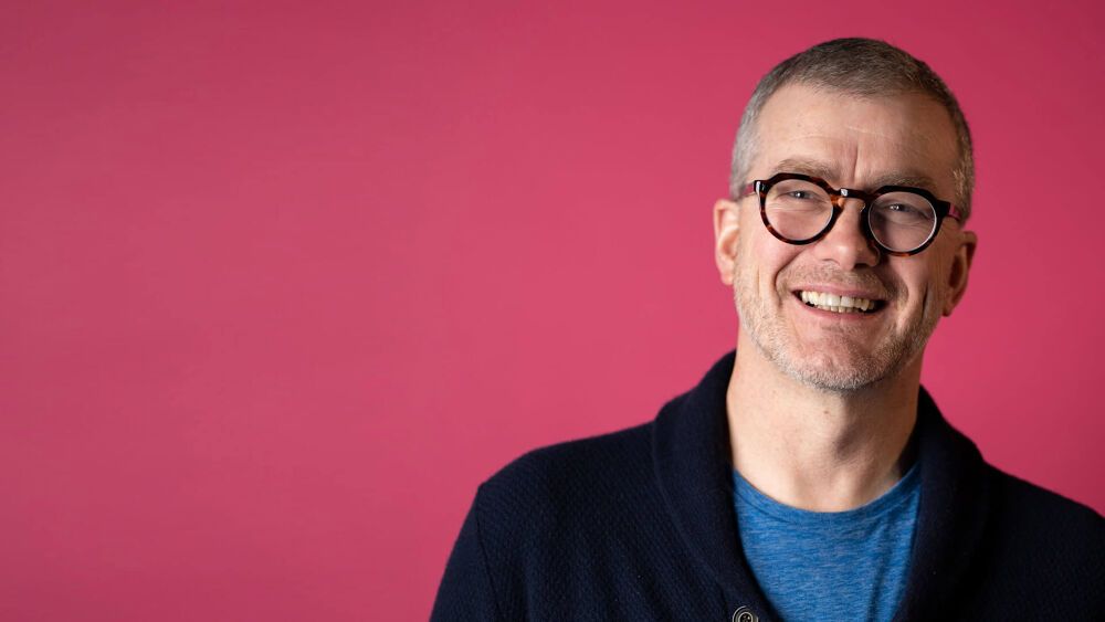 Atom bank CEO, Mark Mullen, wearing glasses and a black cardigan stood in front of a pink background, smiling