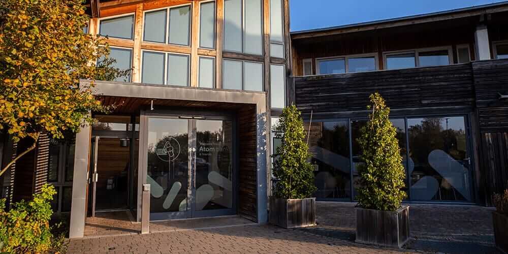 The front door of the Atom bank HQ, the Rivergreen centre, during the end of summer. The sun is setting and there are green trees turning to yellow