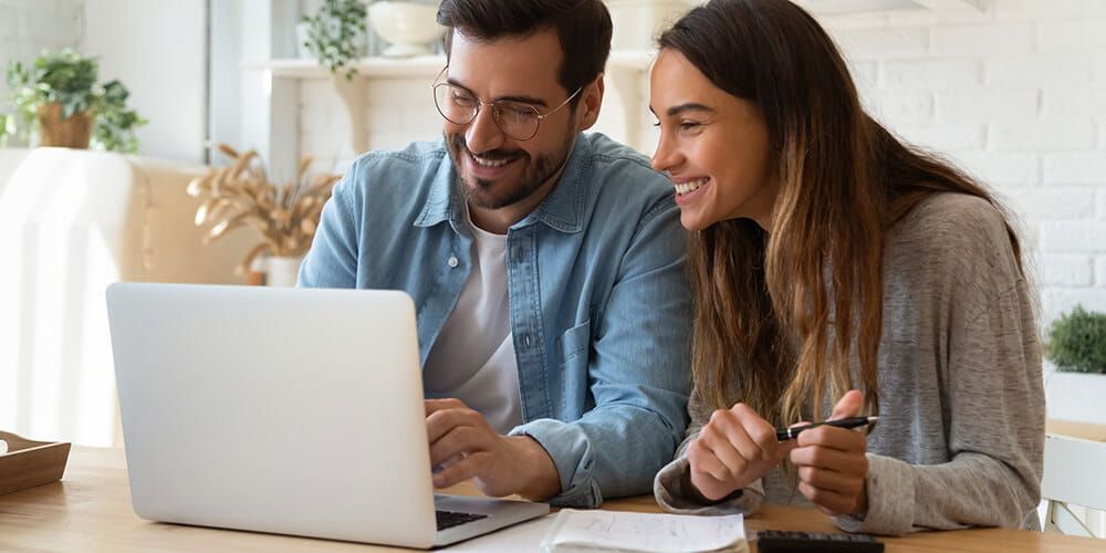 A young couple sitting at the table together smiling at a laptop whilst then organise their finances