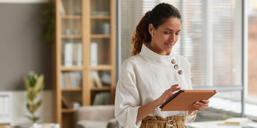 Young woman wearing business casual attire is holding an ipad and wearing wireless earphones whilst smiling in her living room