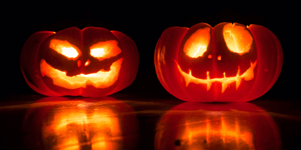 Two illuminated carved pumpkins on a shiney table top casting reflections in dark room