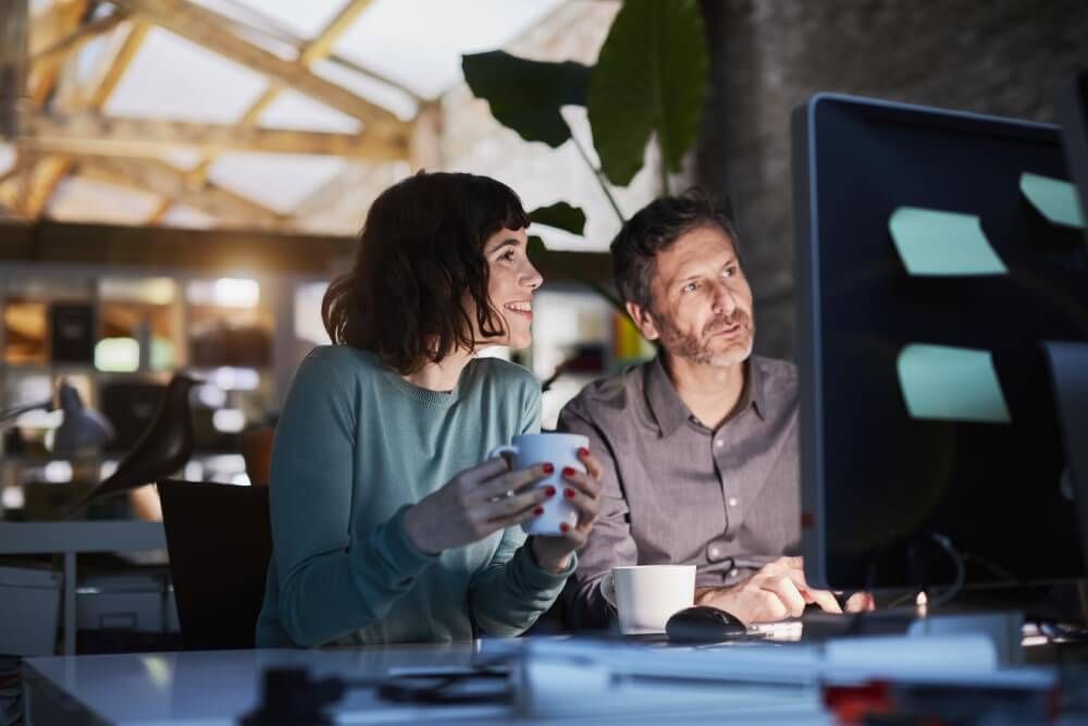 Man and woman in a dimly lit office looking at a bright computer screen whilst drinking from mugs