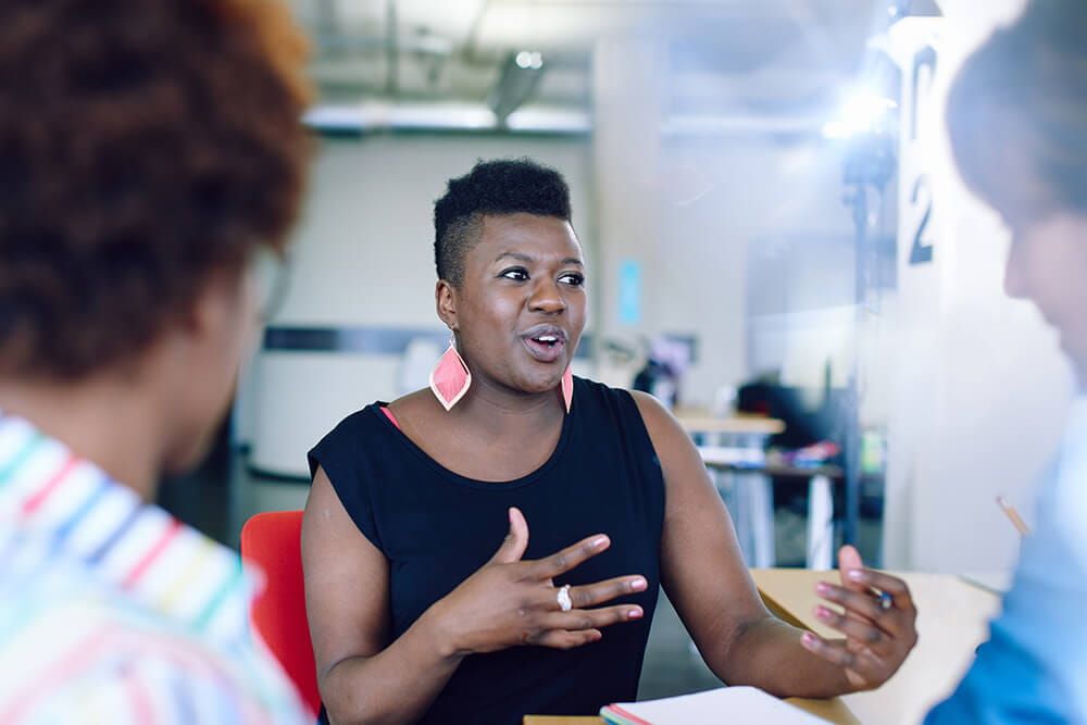 Woman dressed in business casual attire is talking her ideas through with two colleuges whilst holding a pen with her notebook on the table infront of her