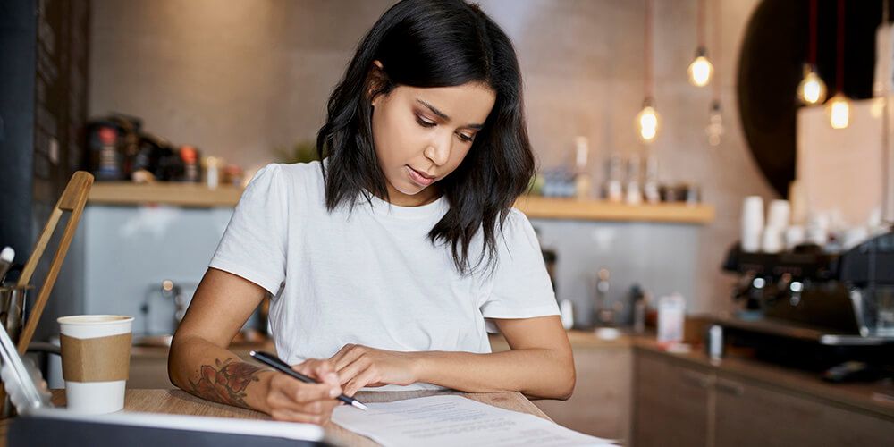 A woman sitting at a table, pen in hand, looking at paperwork