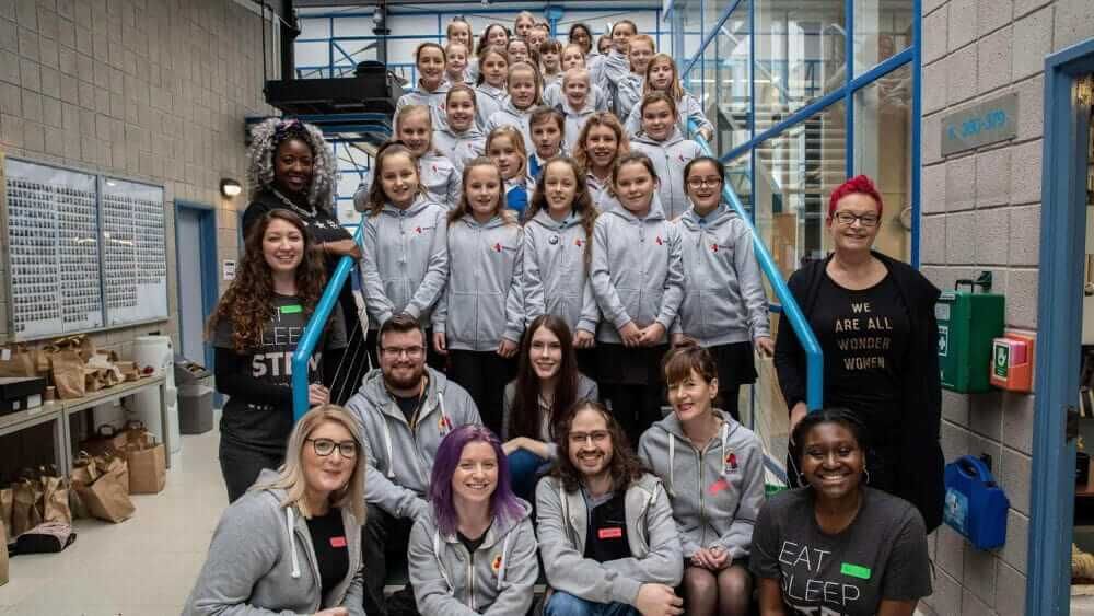 A class of young girls wearing Atom bank hoodies, surrounded by coding professionals