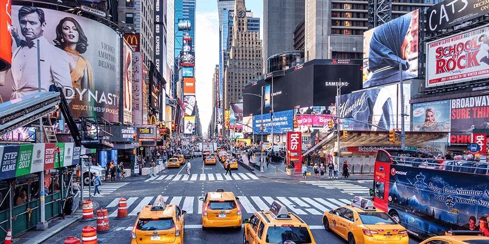 Time square in New York City, there are yellow cabs driving down the street towards the iconic billboards. It is very busy