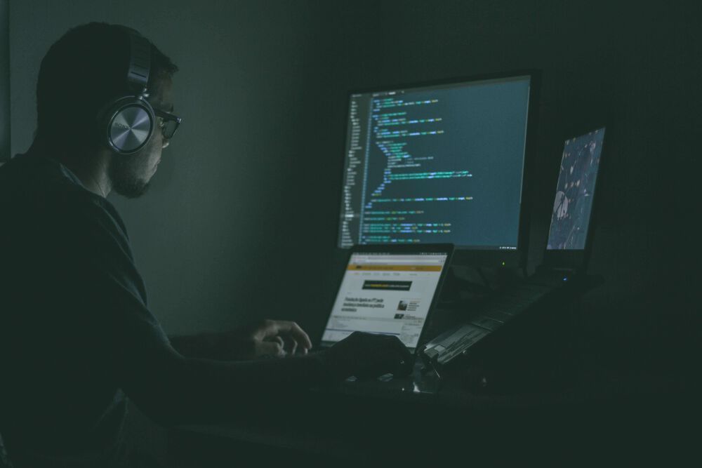 Man sits in very dark room, lit only by dull computer monitors. He is wearing headphones and working across 3 different monitors. He appears to be coding
