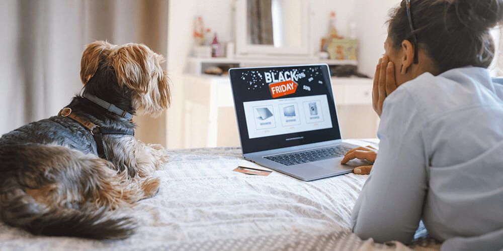 Woman lays with her dog on her bed whilst looking up black friday deals on her laptop. Her credit card is sat next to her