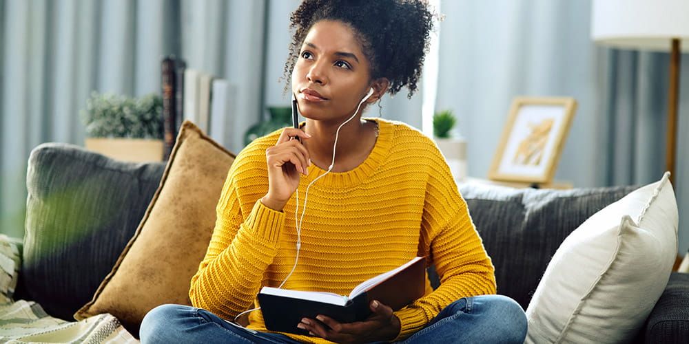 Woman wearing yellow sweater, sat on her sofa listening to music whilst journalling. She looks deep in thought