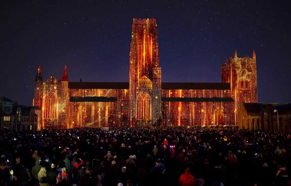 Durham Cathedral lit up in bright gold and orange lights for the Lumiere event, a huge crowd of people stand in the dark garden to look under the night sky