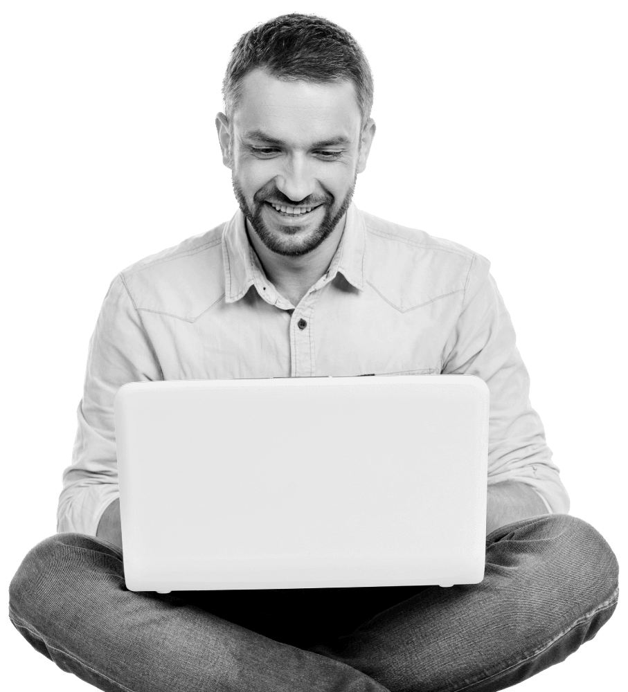 Man sat cross legged with a laptop on his knees, wearing a blue tshirt, smiling