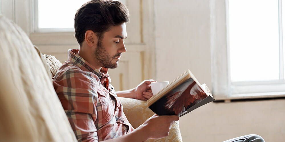 Man sat on a sofa holding a mug, reading a book. He has a concentrated look on his face