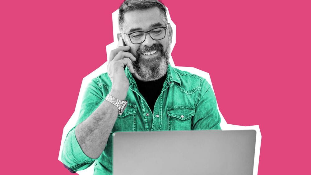 Older man with a beared and green button up shirt, working on his laptop whilst also on the phone