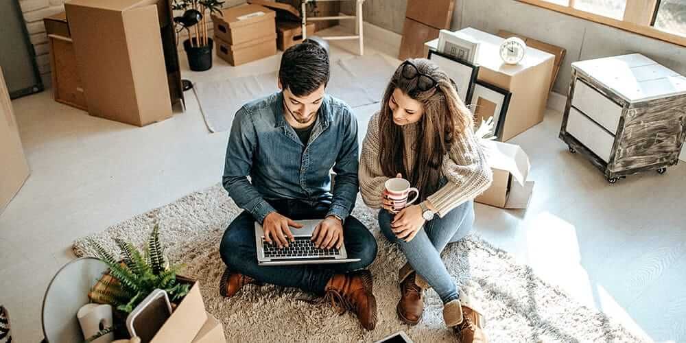 Man and woman sit on the floor on a rug looking together at the laptop sat on the man's knees. They are surrounded by cardboard boxes