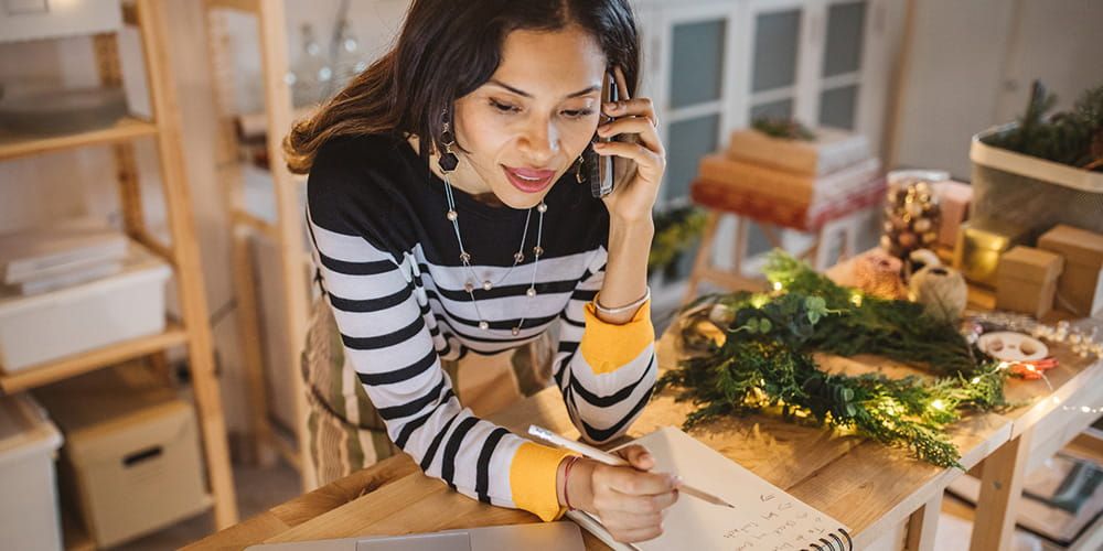 Woman is on the phone with a pencil in her other hand making notes. Next to her is a Christmas wreath