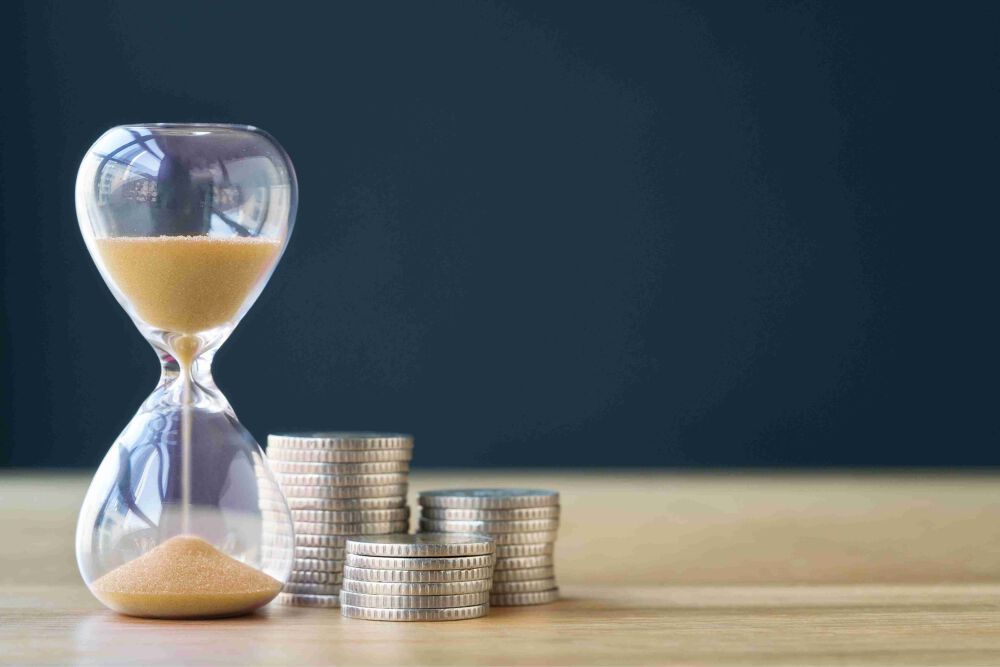 Hour glass with yellow sand falling through it is stood next to a small pile of silver coins