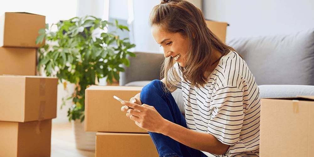 Woman sat smiling at her phone in her living room with moving boxes surrounding her