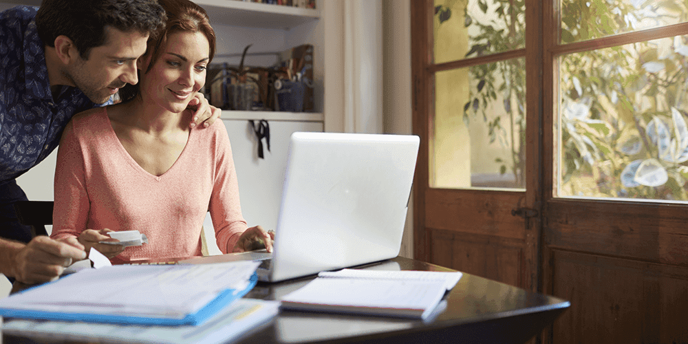 Woman with peach top is sat at her desk, smiling looking at a laptop whilst a man in a blue shirt stood next to her bends to look at the laptop, with his arm around her