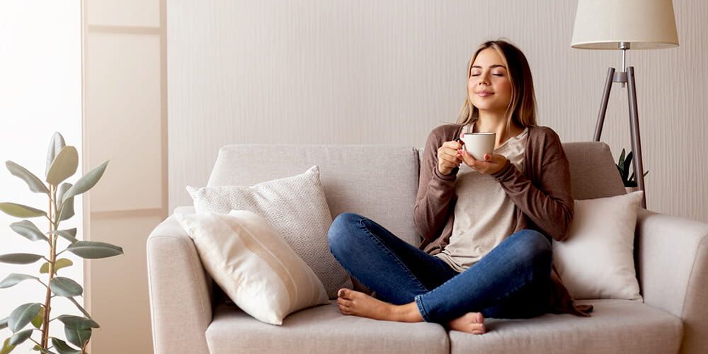Woman sat cross legged on a sofa. She is holding a mug, closing her eyes and taking a deep breath in whilst smiling