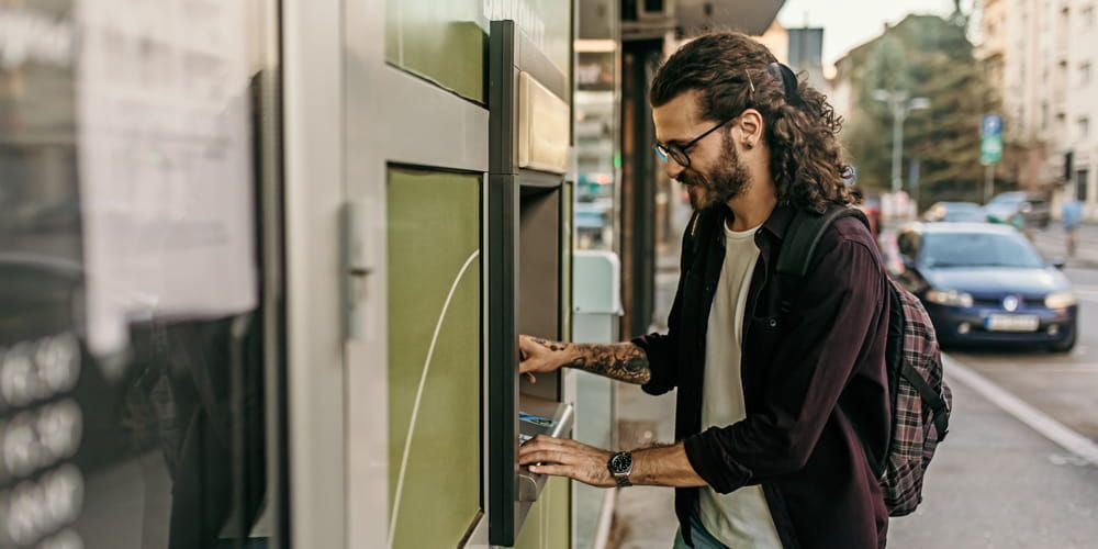 Bearded man with long hair and tattoos smiling whilst using an ATM on a high street