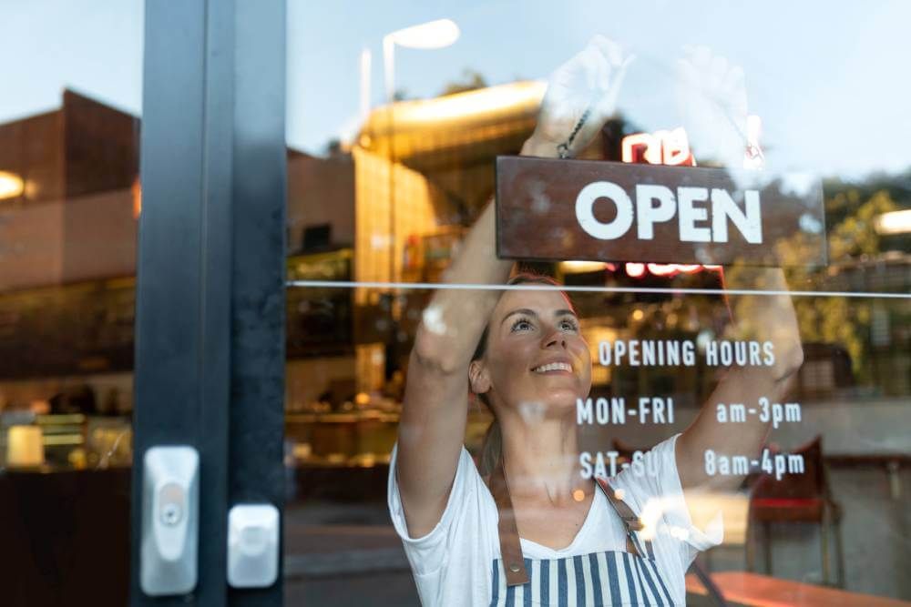 Woman stood in the window of a cafe wearing a striped apron, smiling as she puts the Open sign up
