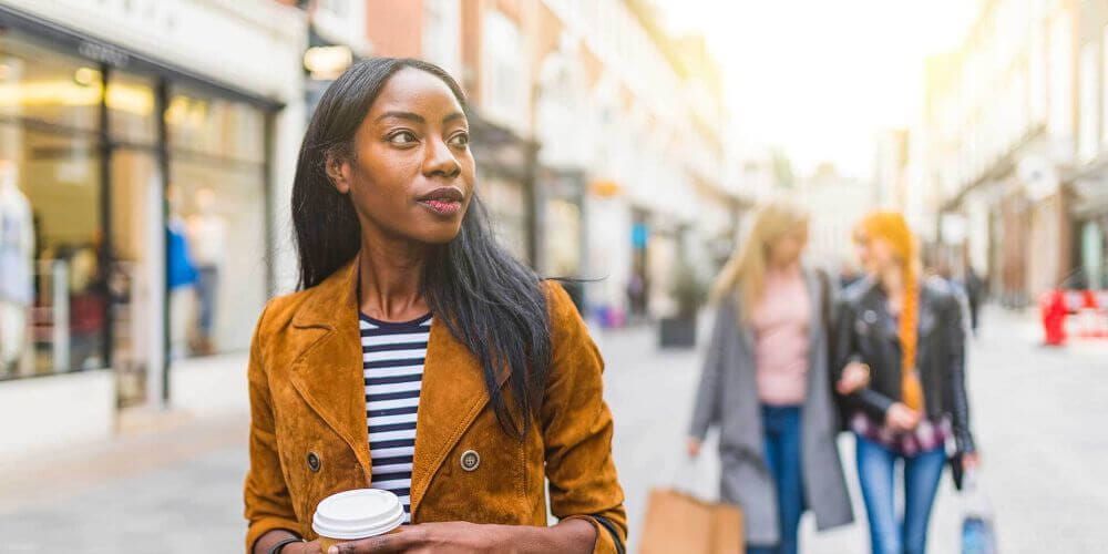 Woman walking down the high street holding a take away coffee cup. In the background two woman walk behind her with shopping bags, talking to each other