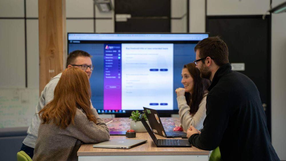 Four Atom bank employees sat around a table with their laptops, working on a presenation together on a larger screen behind them