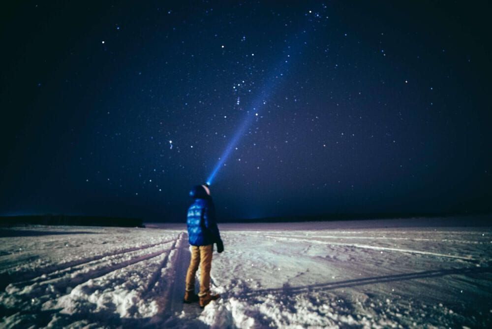 Man wearing blue puffer jacket, stood in the snow, shining a torch into the sky to see a night sky full of stars