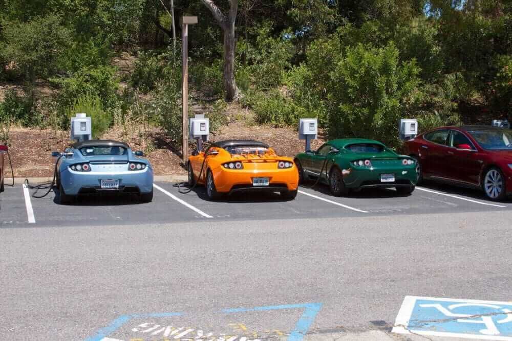 Three sports cars in different colours all parked side by side at a car park with electric chargers plugged into them