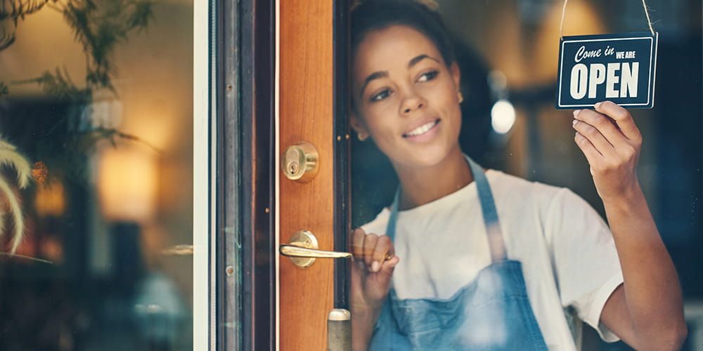 Young woman wearing an apron and smiling is turning a cafe's door sign to Open