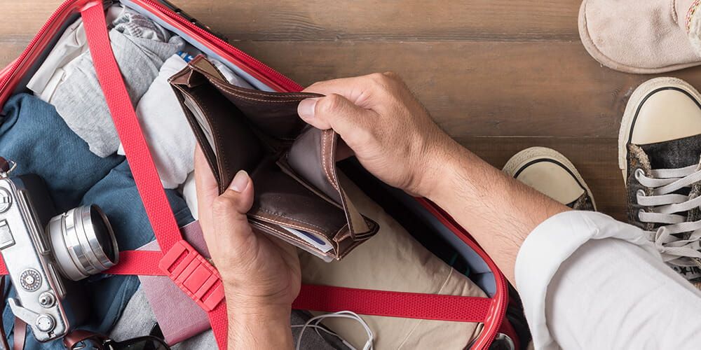 Looking down on a fully packed red suitcase with a camera sat on top, a man is holding open his leather wallet to find it empty