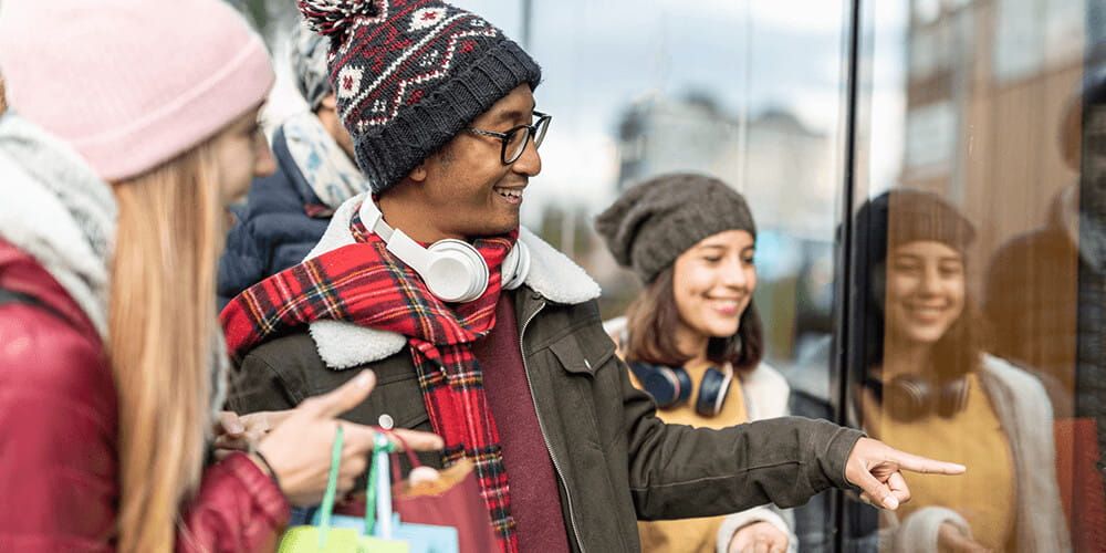 Friends walk point into a shop window dressed in winter clothing. They are smiling and shopping together