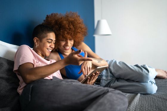 Two woman, one with curly hair and another with very short hair, sat on a bed together looking at an ipad and smiling together