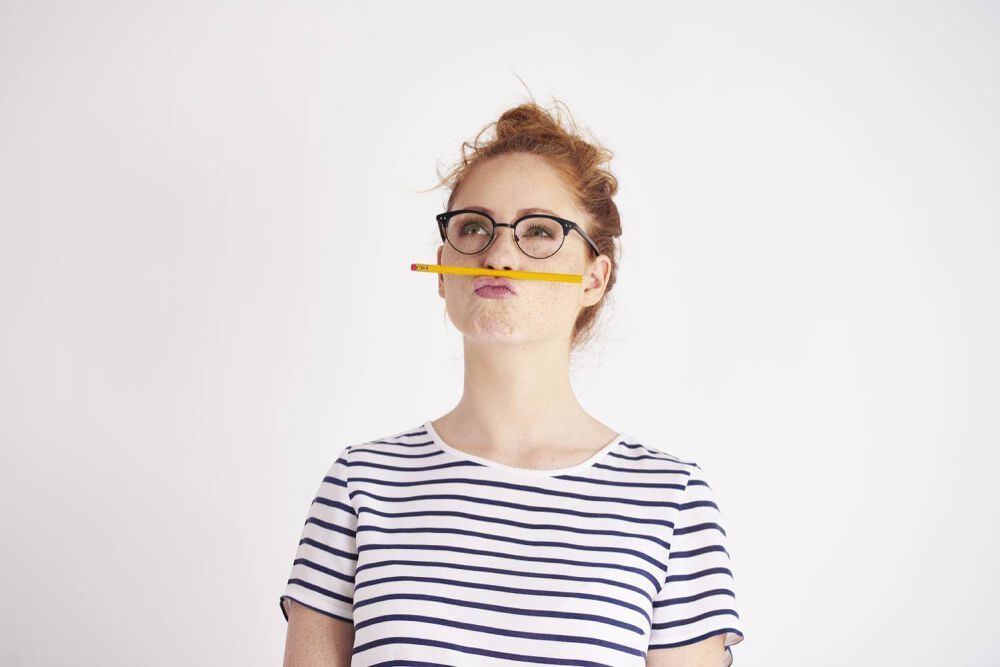 Woman with red hair in a bun, a stripped tshirt and glasses balancing a pencil between her top lip and her nose