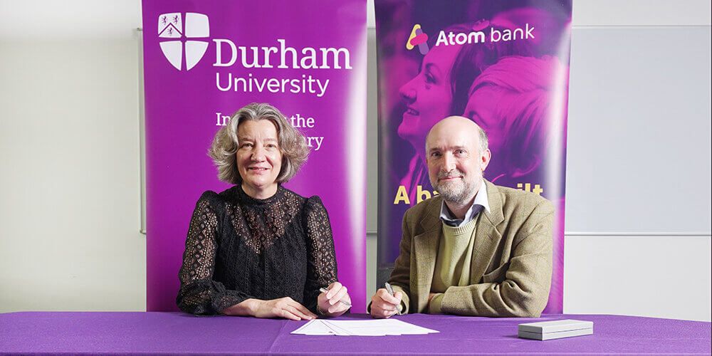 Two people sitting together holding pens about to sign a document, behind them are banners for Durham University and Atom Bank