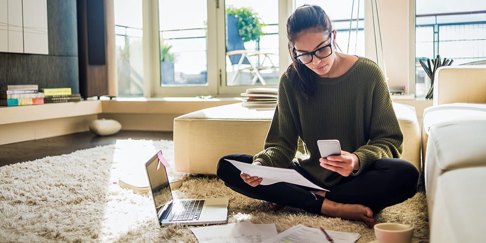 Woman wearing glasses sat crossed legged on the floor of a living room, reading her phone and holding a piece of paper displaying her credit score